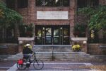 A person rides a bicycle in front of the entrance of The Wharton School''s Steinberg Hall-Dietrich Hall, with brickwork and potted plants.