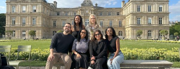 A small group of people posing in front of a grand historic building with a well-manicured garden, under a blue sky with scattered clouds.