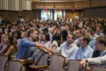 A large group of people seated in an auditorium, with two individuals in the foreground shaking hands, suggesting a social or academic event.