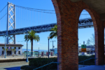 A view of the Bay Bridge in San Francisco framed by brick arches in the foreground. Palm trees line the street near a white building, with a clear blue sky above.