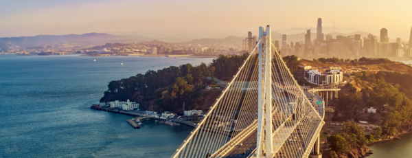 A suspension bridge leading to a city skyline with skyscrapers, seen at sunset. Water and a green, hilly area are visible on the left side. 